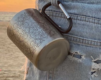 Pode incluir: Uma caneca de viagem de aço inoxidável com um padrão geométrico, presa a um mosquetão. A caneca está pendurada no bolso de trás de um par de calças de ganga azuis.