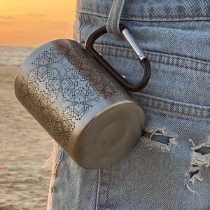Op de afbeelding: Een roestvrijstalen reistumbler met een geometrisch patroon, bevestigd aan een karabijnhaak. De tumbler hangt aan de achterzak van een blauwe spijkerbroek.