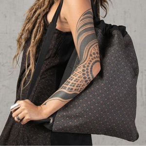 May include: A large, patterned tote bag with a drawstring closure. The bag features a repeating geometric design in shades of gray and brown. The bag has a black strap and a silver grommet detail. The bag is carried over the shoulder.