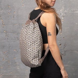 Puede incluir: Una mochila marrón de cordón con un estampado floral blanco y amarillo. La mochila la lleva una persona con el pelo corto y castaño.