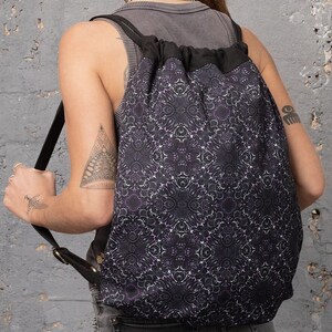 Puede incluir: Una mochila negra de cord&oacute;n con un patr&oacute;n geom&eacute;trico morado y blanco. La mochila tiene correas ajustables y un cierre de cord&oacute;n negro.