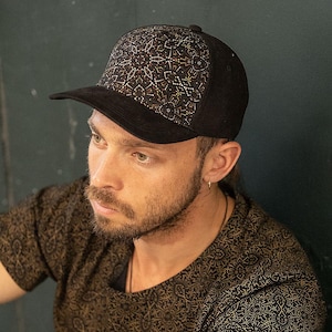 May include: Black baseball cap with a patterned design on the front panel. The cap features a curved brim and a structured crown. The design on the front panel incorporates gold and white accents. The cap is worn with a patterned shirt.