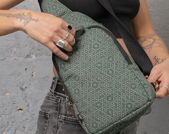 May include: Green and white patterned sling bag with a black adjustable strap and a gold-colored buckle. The bag has a zippered main compartment and a small front pocket. The bag is made of a durable fabric.