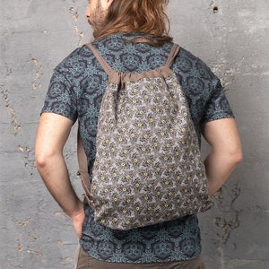 Puede incluir: Una mochila marrón de cordón con un patrón de plumas de pavo real. La mochila tiene correas ajustables y un cierre de cordón.