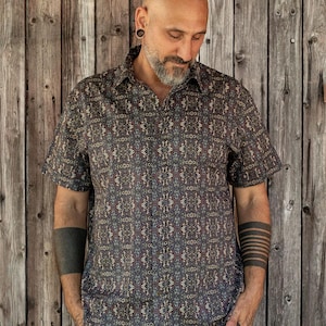 May include: A short-sleeved button-down shirt featuring a dark, intricate, repeating geometric pattern in shades of blue, gold, and brown. The shirt has a classic collar, a front button placket, and a straight hemline, providing a structured fit.