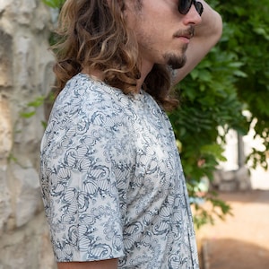 May include: A man wearing a white short-sleeved shirt with a blue and gray abstract pattern. He is wearing sunglasses and has long, wavy hair.