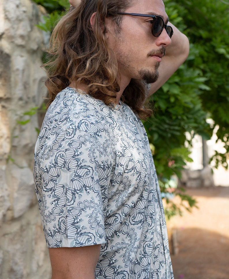 May include: A man wearing a white short-sleeved shirt with a blue and gray abstract pattern. He is wearing sunglasses and has long, wavy hair.