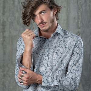 May include: A man wearing a light blue button-down shirt with a white and blue patterned design. The shirt has a long sleeve and the man is wearing several silver rings on his fingers.