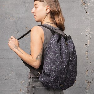 Puede incluir: Una mochila negra de cord&oacute;n con un estampado floral morado y blanco. La mochila tiene una cremallera lateral y correas ajustables.