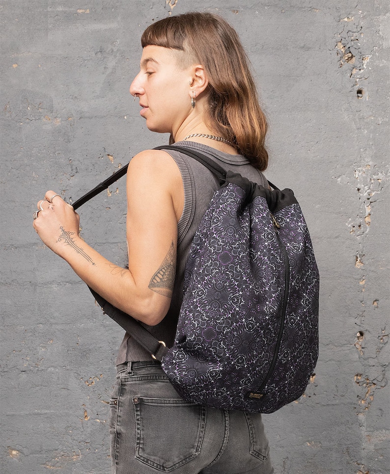 Puede incluir: Una mochila negra de cord&oacute;n con un estampado floral morado y blanco. La mochila tiene una cremallera lateral y correas ajustables.