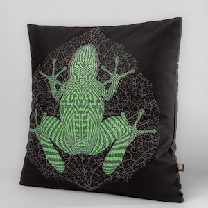 May include: A black cushion featuring a printed green frog. The frog is surrounded by a leaf pattern. The frog has a detailed, geometric design.