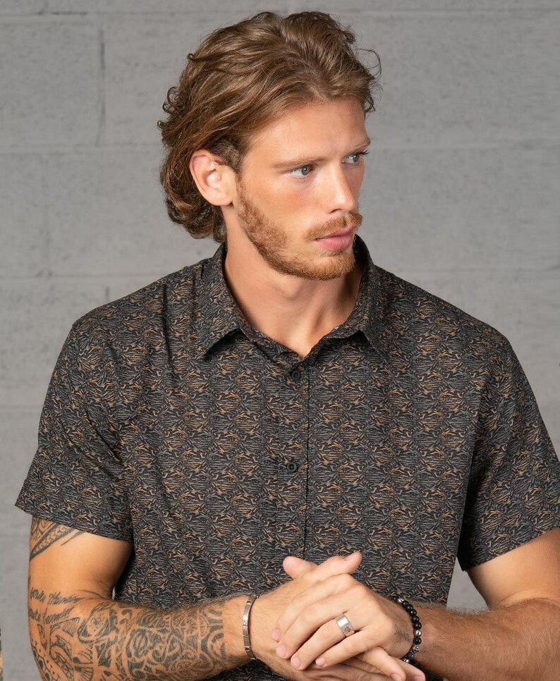 May include: A man wearing a short-sleeved button-down shirt with a brown and black abstract print. He is wearing a silver bracelet and a black beaded bracelet on his left wrist. He is also wearing a silver ring on his right ring finger.