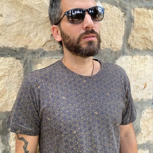 Puede incluir: Un hombre con una camiseta azul oscuro con un patr&oacute;n geom&eacute;trico dorado. Lleva gafas de sol negras y tiene barba.