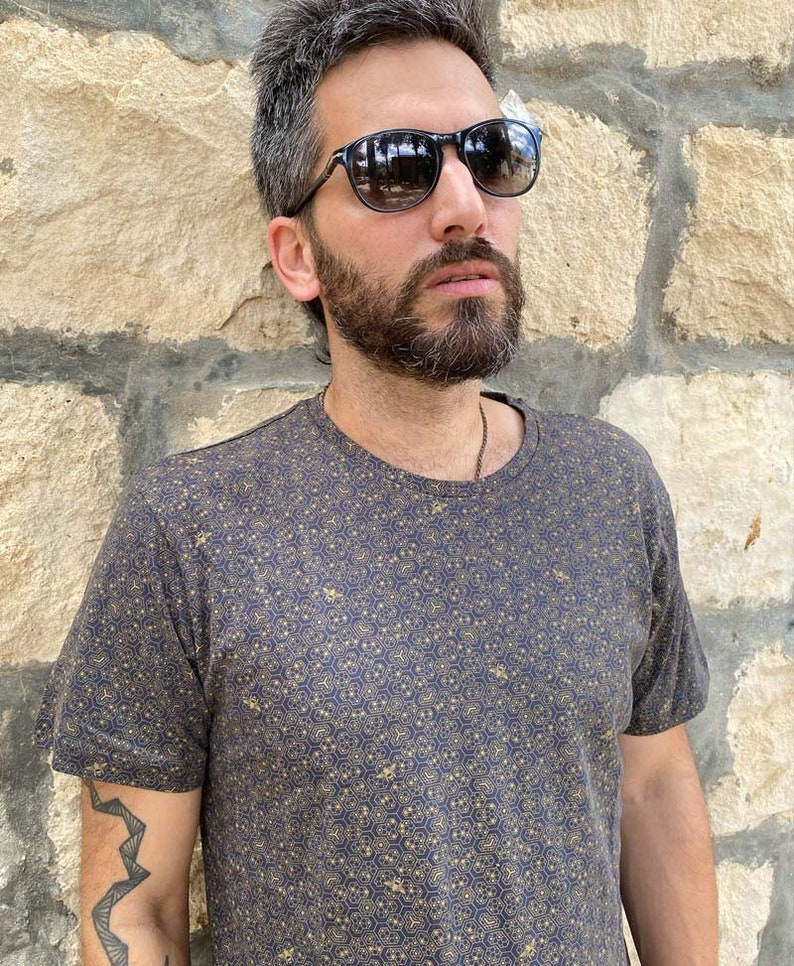 Puede incluir: Un hombre con una camiseta azul oscuro con un patr&oacute;n geom&eacute;trico dorado. Lleva gafas de sol negras y tiene barba.