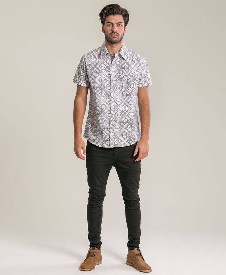 May include: A white short-sleeved button-down shirt with a geometric pattern. The shirt is paired with black skinny jeans and brown leather boots.