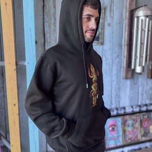 May include: Black hooded sweatshirt with a front pocket and a drawstring hood. The sweatshirt features a gold and orange graphic design on the front. The person is wearing the sweatshirt with their hands in the front pocket.