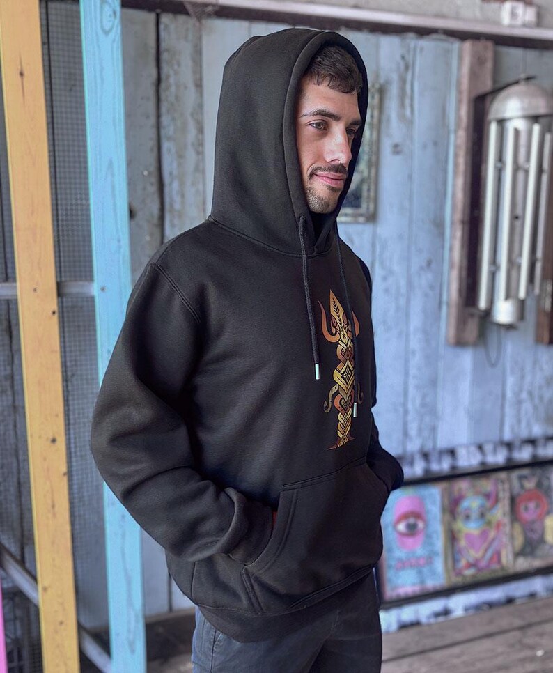 May include: Black hooded sweatshirt with a front pocket and a drawstring hood. The sweatshirt features a gold and orange graphic design on the front. The person is wearing the sweatshirt with their hands in the front pocket.