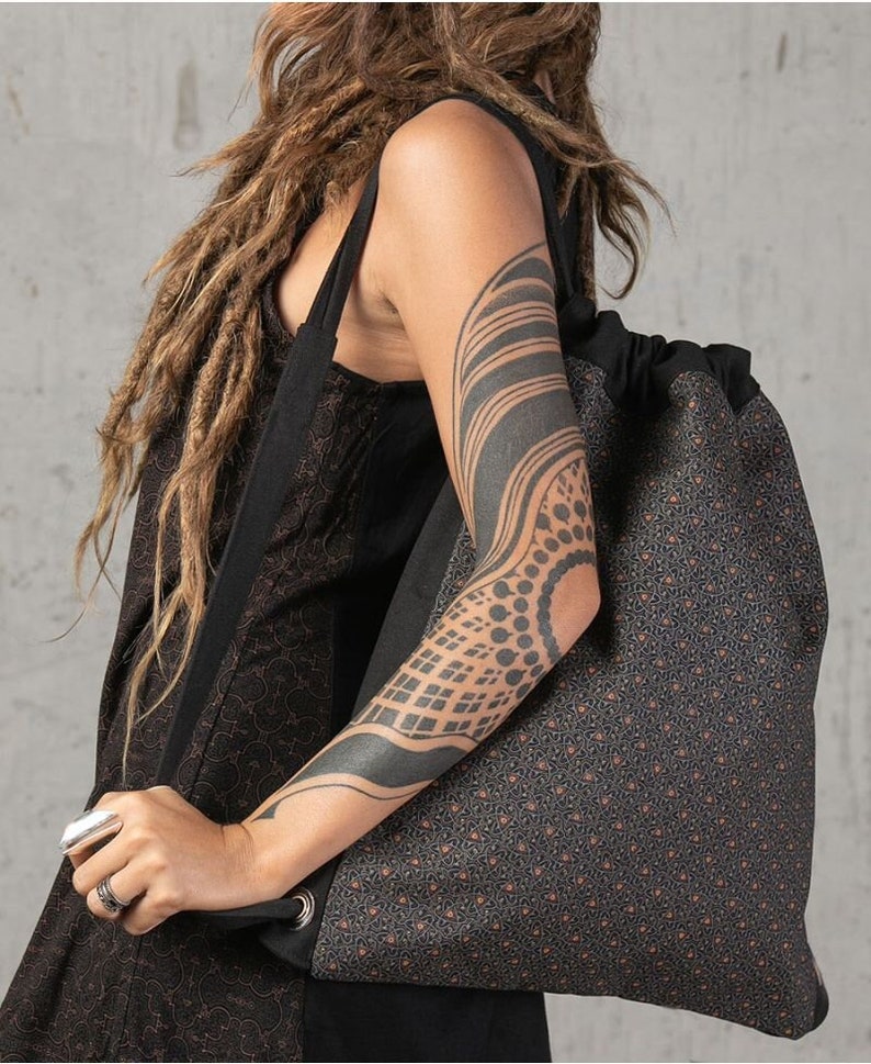 May include: A large, patterned tote bag with a drawstring closure. The bag features a repeating geometric design in shades of gray and brown. The bag has a black strap and a silver grommet detail. The bag is carried over the shoulder.