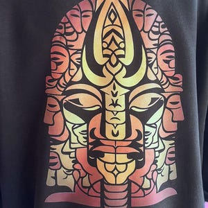 May include: Dark gray sweatshirt featuring a vibrant, stylized graphic of a face composed of multiple faces. The design uses a gradient of orange, yellow, and red hues. The graphic is centered on the front of the shirt.