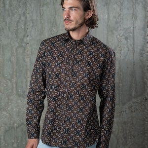 May include: A long-sleeved button-down shirt with a brown background and a repeating pattern of white, navy, and purple floral shapes. The shirt is worn with light blue jeans.