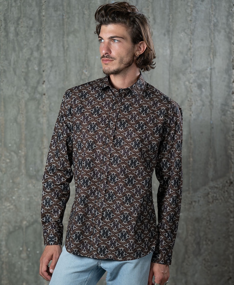May include: A long-sleeved button-down shirt with a brown background and a repeating pattern of white, navy, and purple floral shapes. The shirt is worn with light blue jeans.