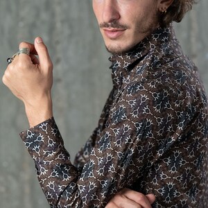 May include: A brown button-up shirt with a repeating white and navy blue floral pattern. The long-sleeved shirt has a high collar and is worn by a person. The shirt is made of a soft, comfortable material.