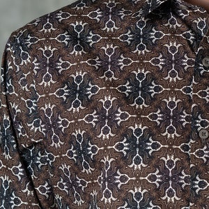 May include: Close-up of a button-down shirt with a repeating geometric pattern in brown, white, and navy blue. The shirt's fabric has a subtle sheen, and the collar and buttons are visible.