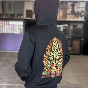 May include: Black hooded sweatshirt with a colorful graphic on the back. The graphic is a stylized face with orange, yellow, and red tones. The sweatshirt has a drawstring hood and long sleeves.