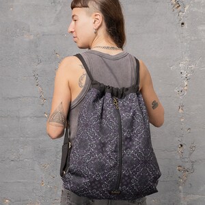 Puede incluir: Una mochila negra de cord&oacute;n con un bolsillo con cremallera y un patr&oacute;n de mandala blanco y morado. La mochila la lleva una persona con un tatuaje en el brazo.