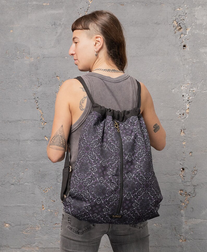Puede incluir: Una mochila negra de cord&oacute;n con un bolsillo con cremallera y un patr&oacute;n de mandala blanco y morado. La mochila la lleva una persona con un tatuaje en el brazo.