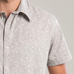 Puede incluir: Una camisa de manga corta de color gris claro con botones y un patrón geométrico. La camisa tiene un bolsillo en el lado izquierdo.