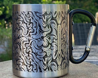 Pode incluir: Caneca de viagem em aço inoxidável com uma alça preta e um padrão gravado. A caneca tem uma alça estilo mosquetão e o endereço do site www.seedoflife.shop está impresso na lateral. A caneca está em uma superfície de pedra.