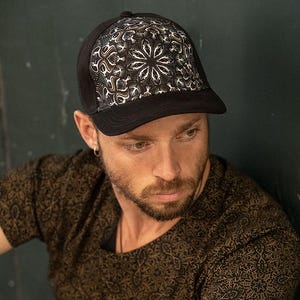 Puede incluir: Una gorra de béisbol negra de malla con un diseño de mandala blanco y dorado. La gorra tiene una visera curvada y una correa ajustable negra.
