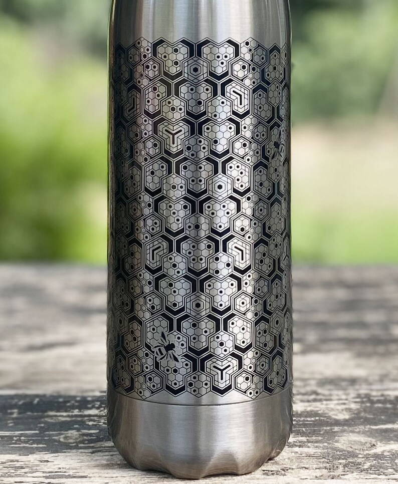 Pode incluir: Garrafa de &aacute;gua de a&ccedil;o inoxid&aacute;vel prateado com um padr&atilde;o de favo de mel gravado. O design inclui uma abelha estilizada. A garrafa tem uma base curva e &eacute; adequada para bebidas.