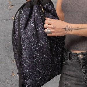 Puede incluir: Una bolsa de cord&oacute;n negro con un dise&ntilde;o estampado en morado y blanco. La bolsa tiene una cremallera y un cord&oacute;n de ajuste negro.
