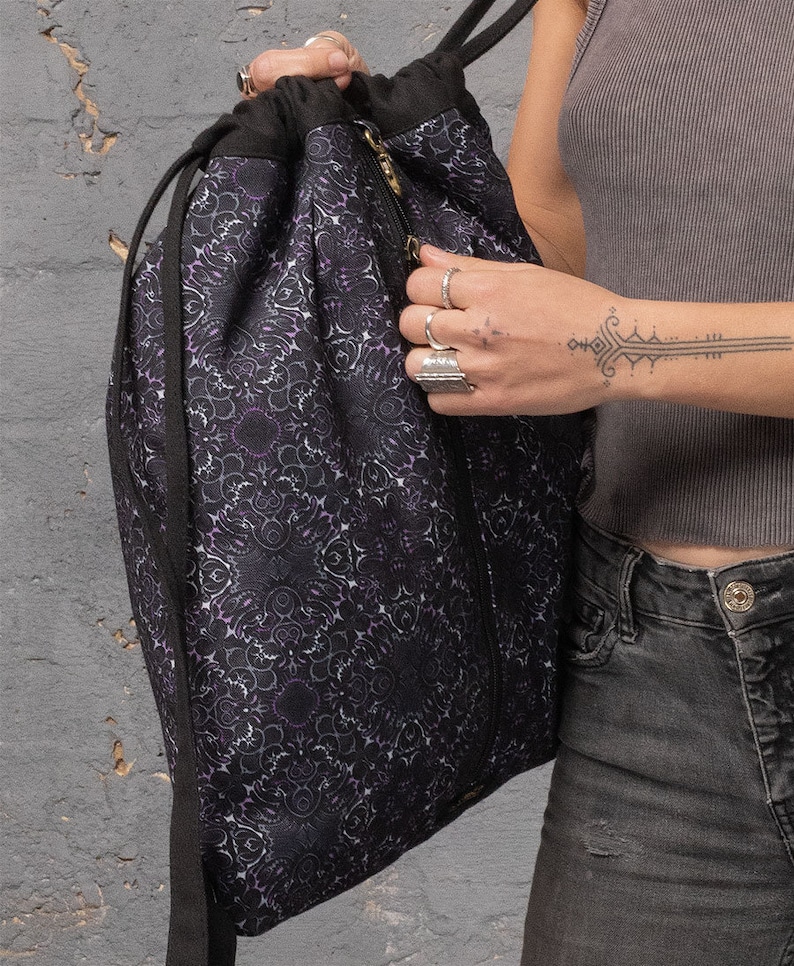 Puede incluir: Una bolsa de cord&oacute;n negro con un dise&ntilde;o estampado en morado y blanco. La bolsa tiene una cremallera y un cord&oacute;n de ajuste negro.
