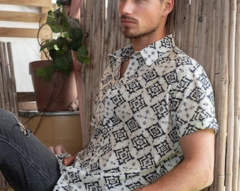 Op de afbeelding: Kort mouwshirt met een zwart-wit geometrisch patroon. Het shirt wordt gecombineerd met een donkerblauwe jeans. Het model zit op een houten bank naast een bamboe hek.