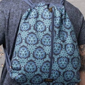 Puede incluir: Una mochila azul de cord&oacute;n con un bolsillo con cremallera y un patr&oacute;n geom&eacute;trico de copo de nieve. La mochila tiene correas ajustables y una peque&ntilde;a etiqueta que dice "SOL SEED OF LIFE".