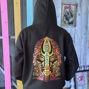 May include: A black hooded sweatshirt with a colorful graphic design on the back. The design features a stylized face with multiple faces and a symmetrical pattern in shades of orange, yellow, and red.