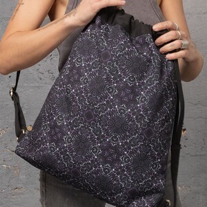 Puede incluir: Una bolsa de cord&oacute;n negro con un patr&oacute;n floral morado y blanco. La bolsa tiene una correa larga y est&aacute; hecha de una tela suave y duradera.