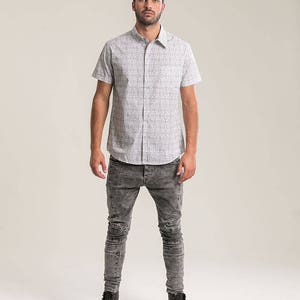 Pode incluir: Um homem vestindo uma camisa branca de manga curta com bot&otilde;es e um padr&atilde;o preto e branco e cal&ccedil;as jeans skinny cinzas. Ele est&aacute; usando botas de couro pretas.
