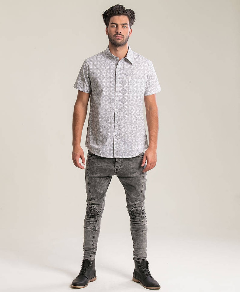 Pode incluir: Um homem vestindo uma camisa branca de manga curta com bot&otilde;es e um padr&atilde;o preto e branco e cal&ccedil;as jeans skinny cinzas. Ele est&aacute; usando botas de couro pretas.