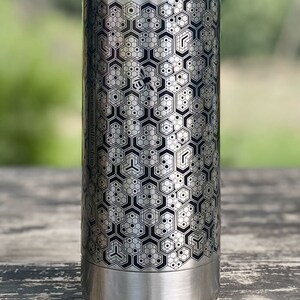 Pode incluir: Garrafa de &aacute;gua de a&ccedil;o inoxid&aacute;vel prateado com um padr&atilde;o geom&eacute;trico de favo de mel e uma abelha. A garrafa tem um design em preto e cinza e o endere&ccedil;o do site www.seedoflife.shop est&aacute; impresso na lateral.