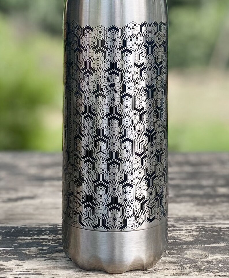 Pode incluir: Garrafa de &aacute;gua de a&ccedil;o inoxid&aacute;vel prateado com um padr&atilde;o geom&eacute;trico de favo de mel e uma abelha. A garrafa tem um design em preto e cinza e o endere&ccedil;o do site www.seedoflife.shop est&aacute; impresso na lateral.