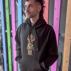 May include: Black hoodie with a front pocket and a hood. The hoodie features a colorful, detailed graphic design on the front. The design is a vertical, symmetrical pattern with orange, yellow, and brown hues.