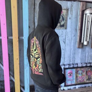 May include: Black hooded sweatshirt with a colorful graphic design on the back. The design features a stylized face with multiple eyes and a mix of orange, yellow, and pink hues. The sweatshirt is worn by a person.