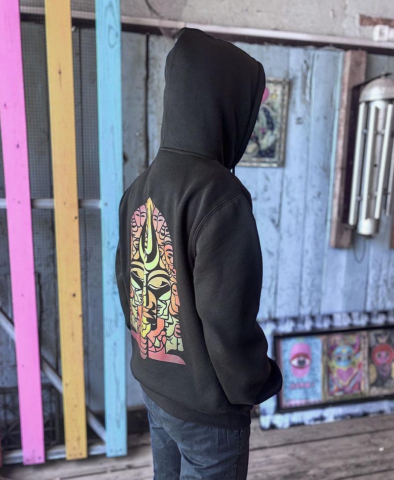 May include: Black hooded sweatshirt with a colorful graphic design on the back. The design features a stylized face with multiple eyes and a mix of orange, yellow, and pink hues. The sweatshirt is worn by a person.