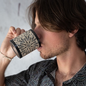 May include: A person drinks from a black and gold ceramic mug with an intricate geometric pattern. The mug is held in the person's right hand.
