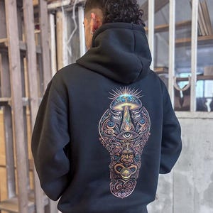 May include: Black hooded sweatshirt with a colorful, intricate graphic on the back. The design features a mushroom, eyes, and other symbolic elements in shades of gold, blue, and pink. The sweatshirt is made of a soft material.