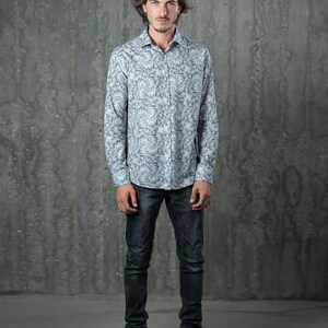 May include: A man wearing a white long-sleeved shirt with a blue and white floral pattern. He is wearing dark blue jeans and green sneakers.